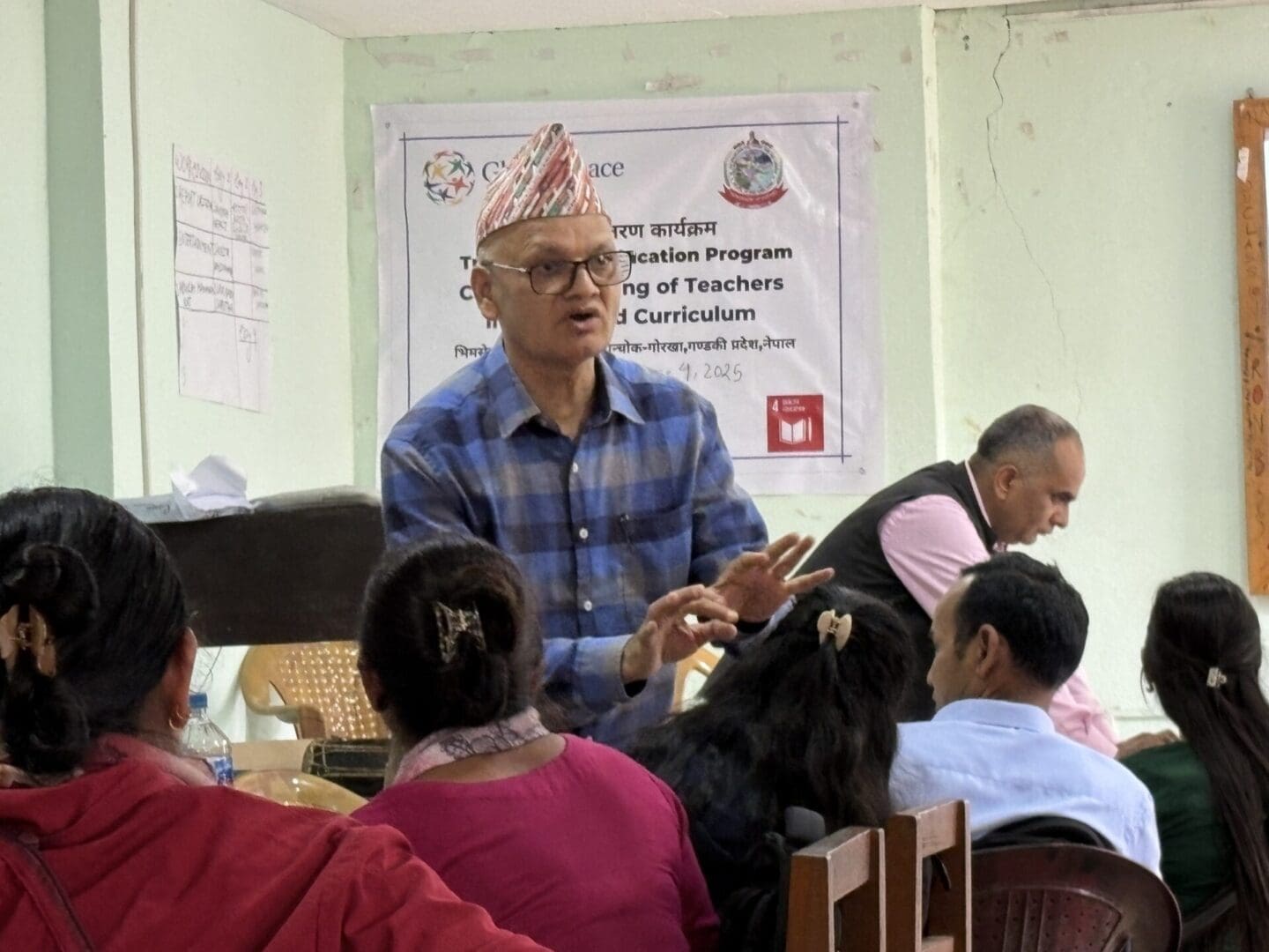 A man wearing a checkered shirt and traditional hat speaks to a group of seated people in a classroom with a banner about capacity building of teachers on the wall behind him, highlighting efforts to strengthen education in Nepal.