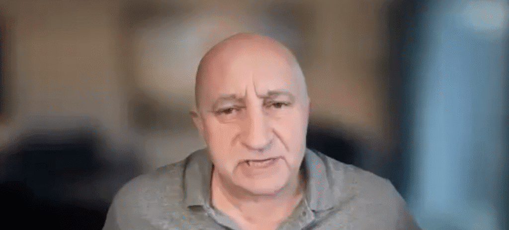 A middle-aged bald man wearing a gray polo shirt looks toward the camera with a neutral expression, sitting indoors with a blurred background—an image often seen in profiles of global network peacebuilders.