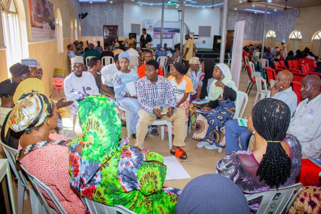 A group of people sit in a circle inside a hall, engaged in a peacebuilding discussion, with others seated in rows in the background at a GPF Nigeria event in Niger State.