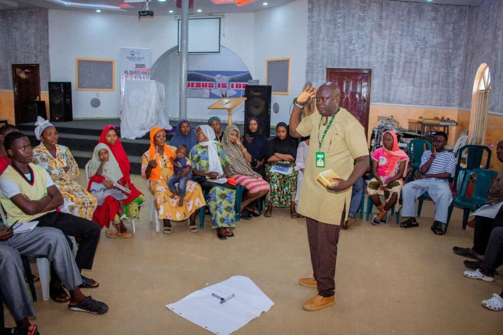 A man stands and speaks to a seated group of men and women arranged in a circle, facilitating a community-based peacebuilding session for GPF Nigeria, with paper and markers on the floor in the center of the room.