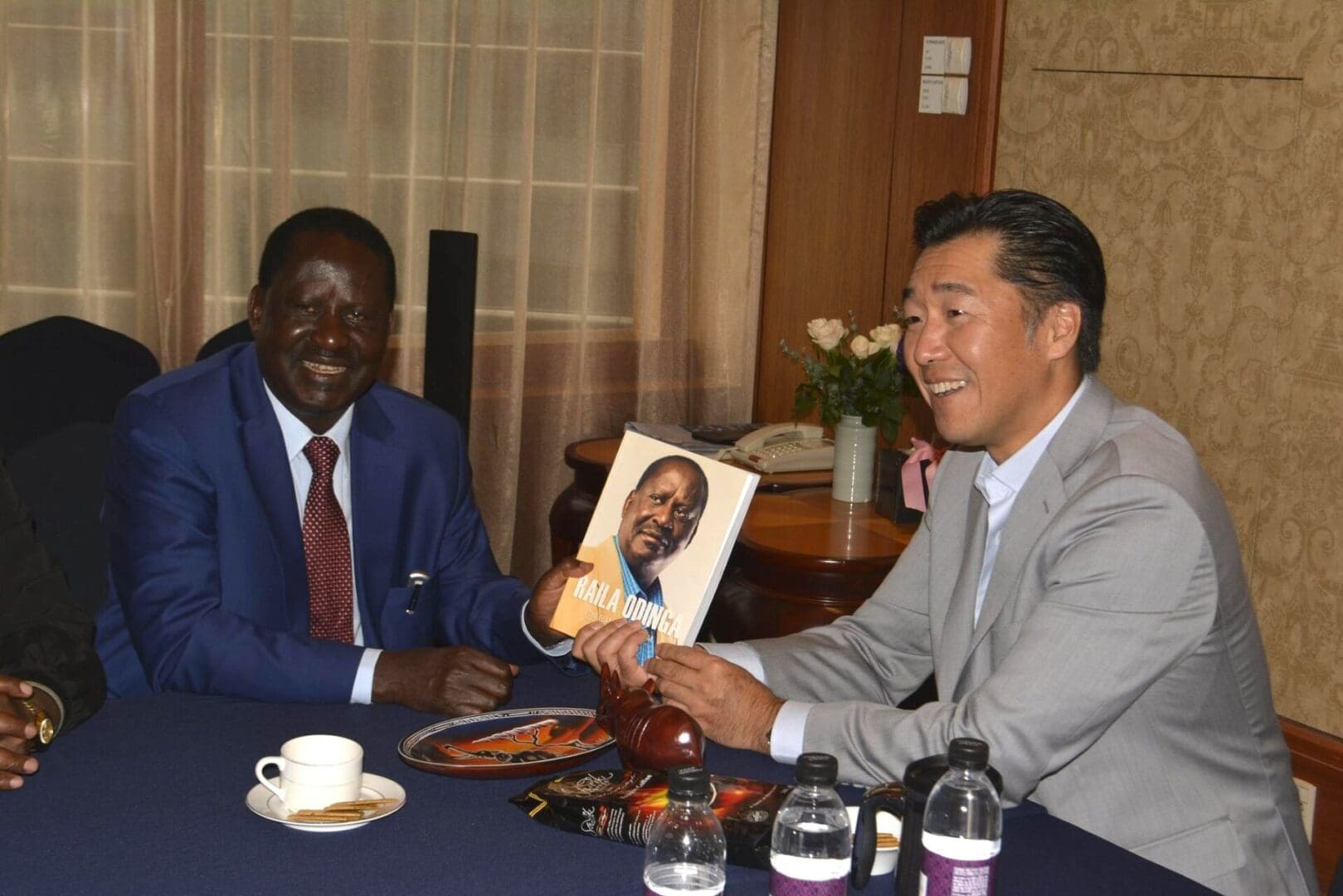 Two men sit at a table; one hands a book with Raila Amolo Odinga's portrait on the cover to the other. Bottled water and plates are on the table, and a vase of flowers is in the background, honoring Rt. Hon. Odinga's legacy.