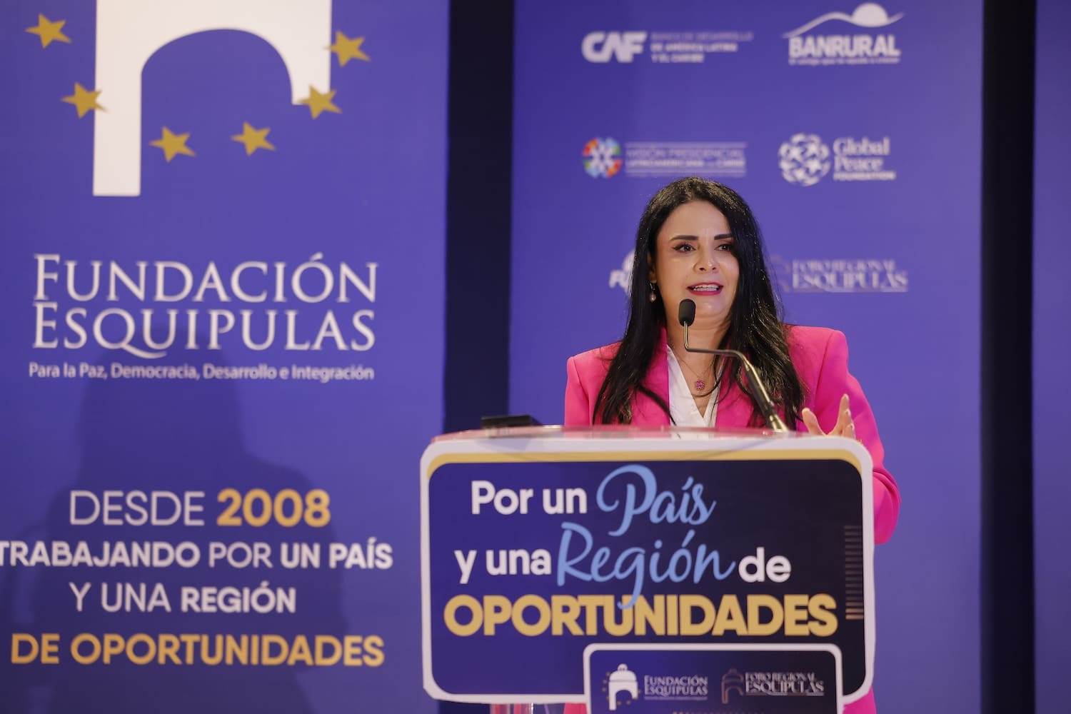 A woman in a pink blazer speaks at a podium during a Fundación Esquipulas event, with banners in the background promoting opportunities, regional development, and strengthening democracy at the Esquipulas Regional Forum 2025.