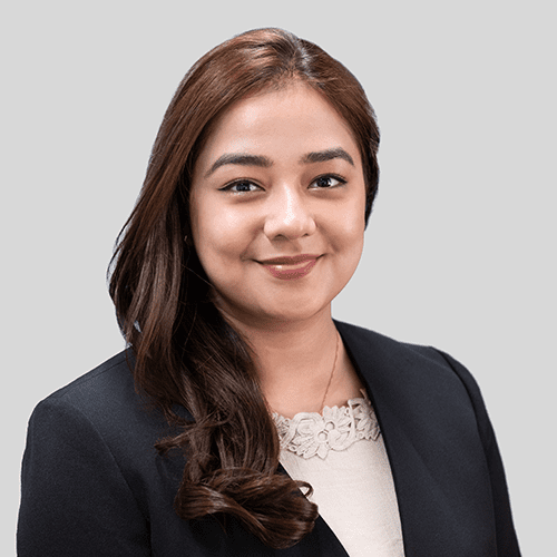 Shraddha KC, a woman with long brown hair wearing a black blazer and beige blouse, smiles at the camera against a plain gray background.