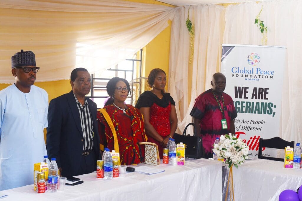 Five people stand behind a table with drinks and flowers at a Global Peace Foundation (GPF) Nigeria event, promoting Peace Education, beside a banner reading