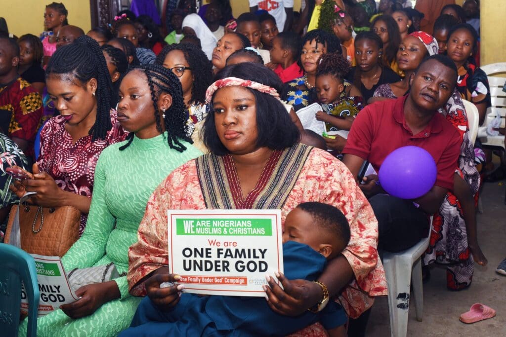 A diverse group of adults and children sit closely together indoors; a woman in front holds a sign reading, “We are one family under God,” promoting peace education amidst insecurity in Nigeria.