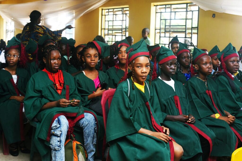 A group of young students wearing green graduation gowns and caps are seated indoors during a Peace Education ceremony, looking toward the camera.