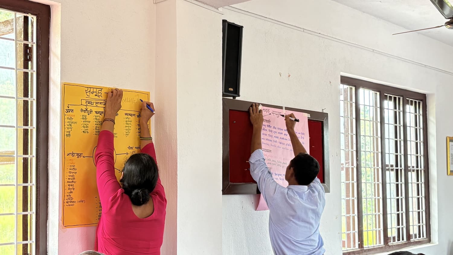 Two people tack large handwritten posters to a white wall in a brightly lit room with barred windows, preparing materials for an Integrated Curriculum workshop focused on capacity building of teachers.