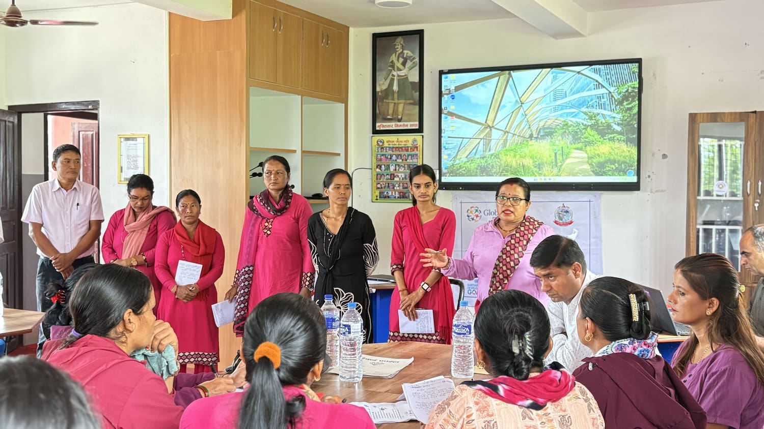 A group of people in a meeting room, with several women standing and speaking while others sit around a table, some holding papers—discussing education in Nepal and the capacity building of teachers. A large screen and posters are visible in the background.