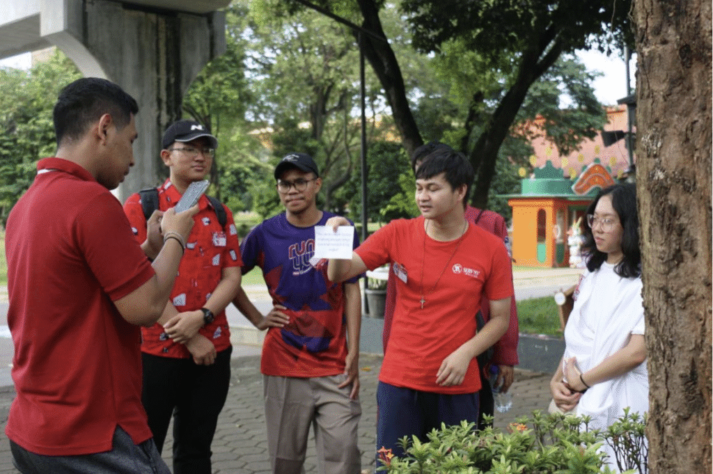 A group of five Youth stand outdoors; one person holds up a note while another takes a photo with a smartphone. Trees and park structures are visible in the background, reflecting GPF Indonesia's commitment to Peacebuilding.