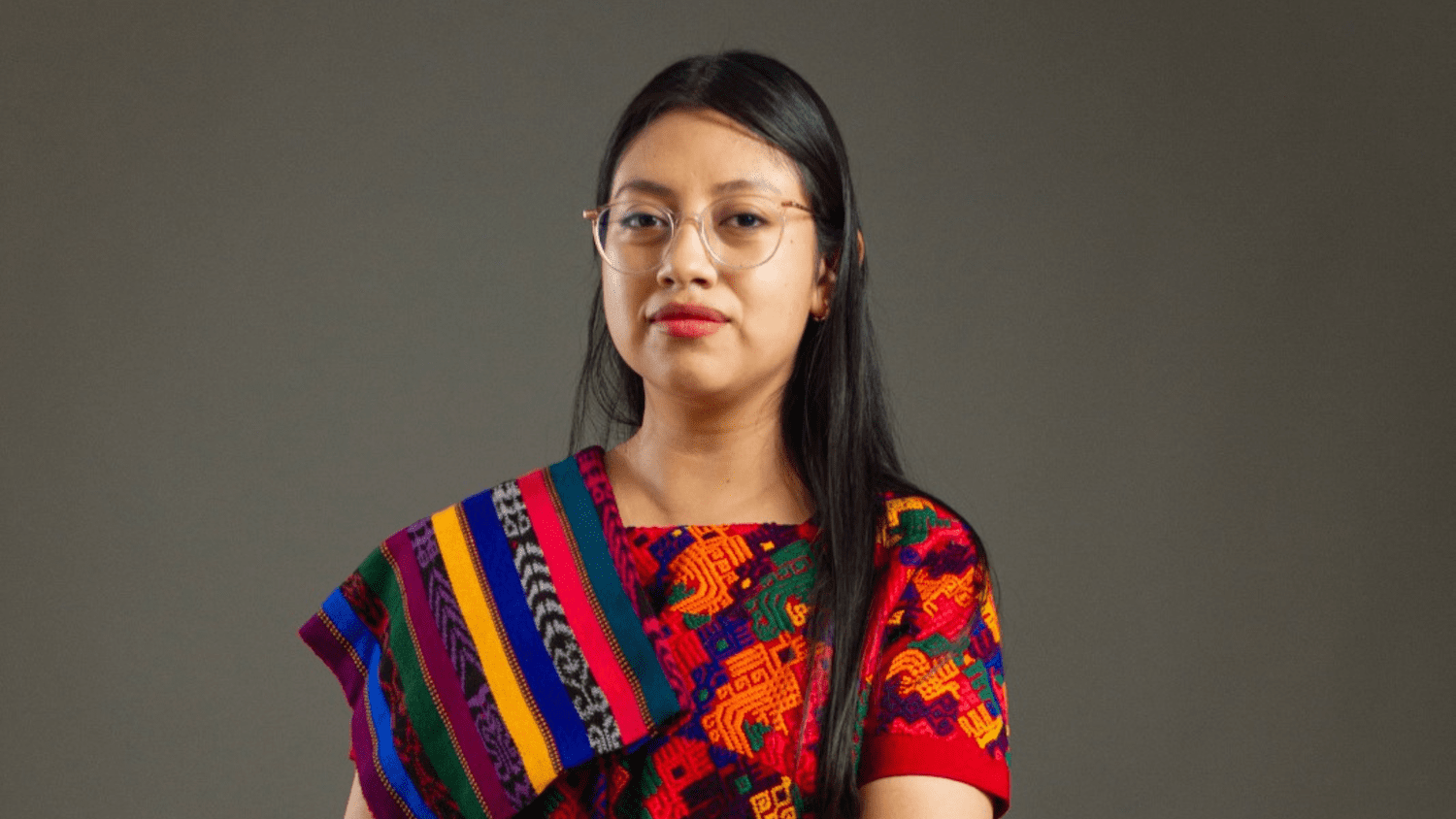 A woman with long black hair and glasses wears a colorful traditional garment with intricate Guatemalan patterns, standing against a plain gray background, evoking the vibrant heritage of Central America.