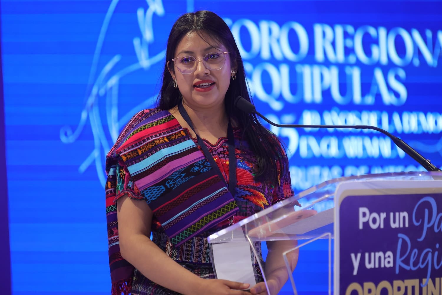 A woman in traditional attire from Guatemala speaks at a podium during the World Food Forum conference, with a blue background displaying event text.