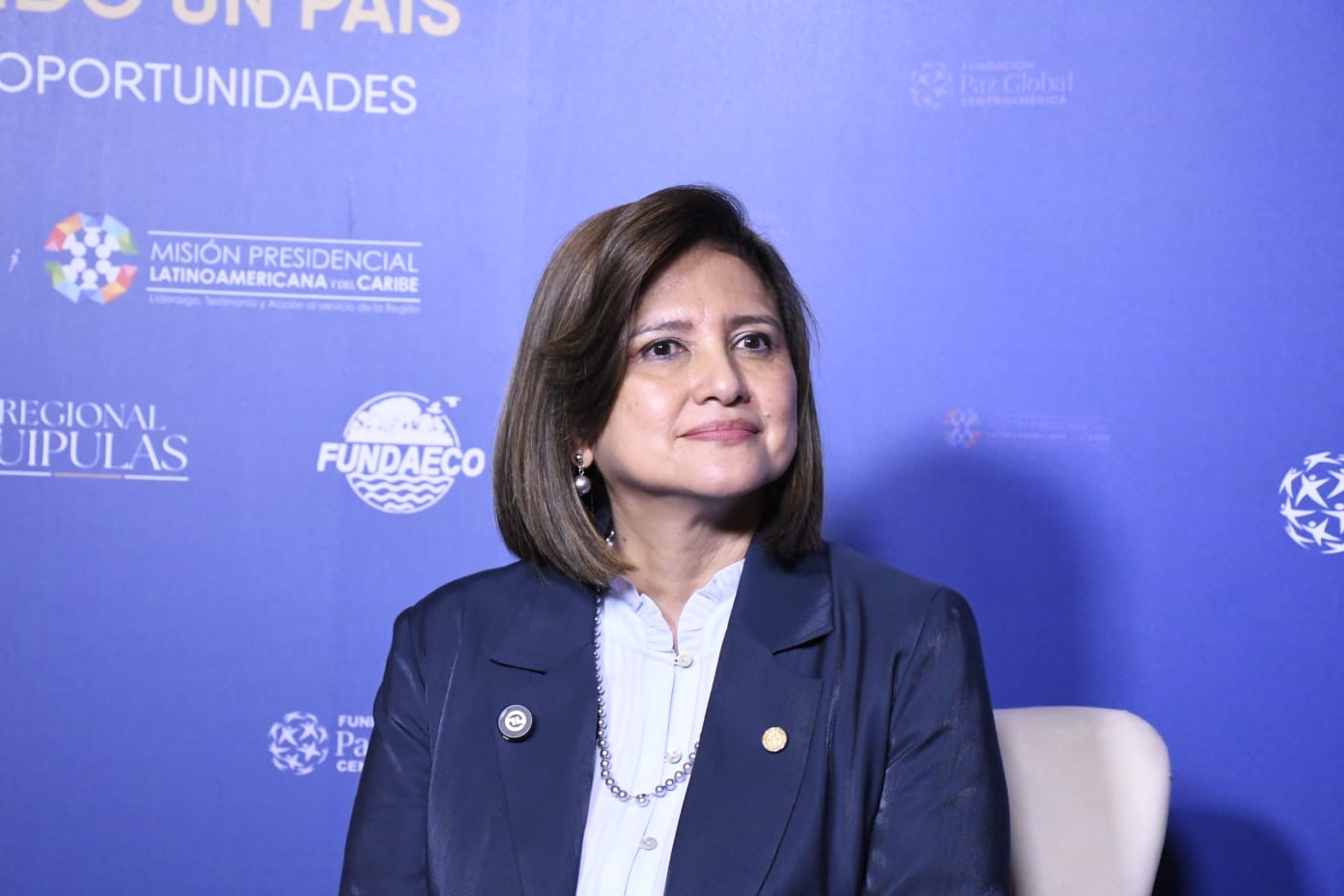 A woman in a navy blazer and white blouse sits in front of a blue backdrop with logos and Spanish text related to democracy and the Esquipulas Regional Forum on Latin American and Caribbean presidential missions.