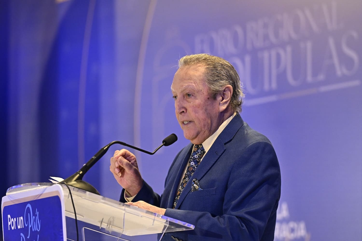 An older man in a blue suit speaks at a podium with a microphone during the Esquipulas Regional Forum, addressing the importance of democracy and regional integration against a striking blue backdrop.
