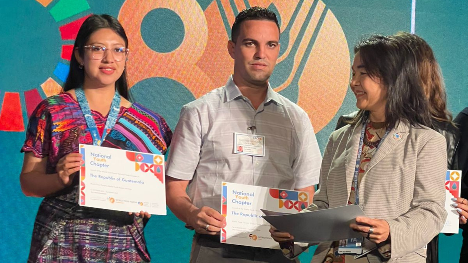 Three people stand together holding certificates at an event, with a colorful backdrop featuring circular graphics and text celebrating Guatemala and Central America, likely as part of the World Food Forum.