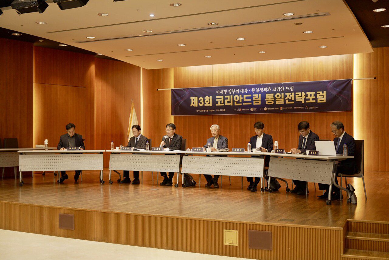 Seven people sit at a long table on a stage during a formal panel discussion, with a blue banner featuring Korean text about the Korean Dream displayed behind them.