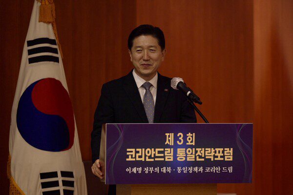 A man in a suit stands at a podium with Korean writing, next to a South Korean flag, discussing national security at the 3rd Korean Dream Unification Strategy Forum.