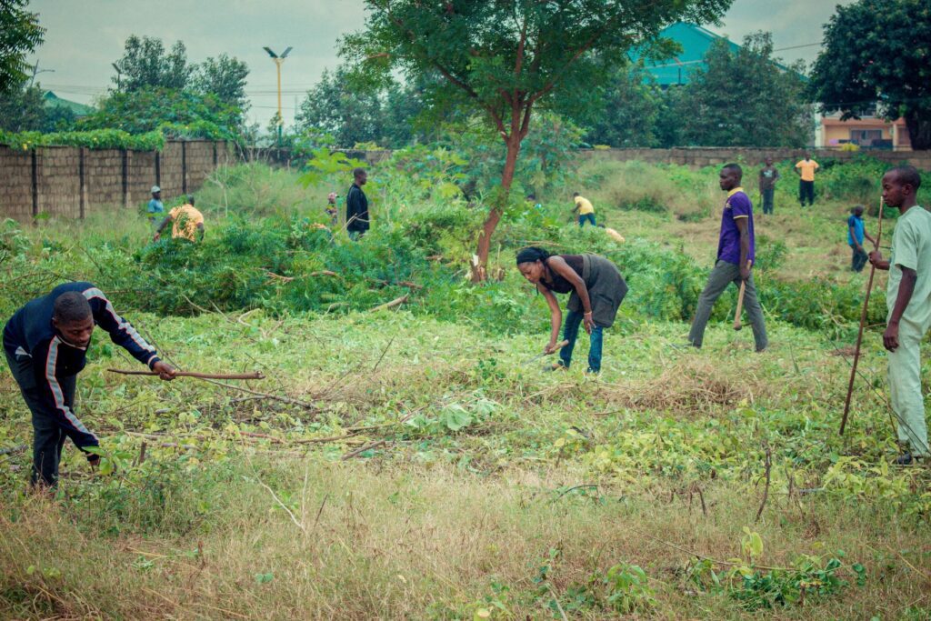 Several people are working outdoors in a grassy field at Hayin Banki, using tools to clear vegetation under cloudy skies. This interfaith service project brings the community together, with trees and distant buildings visible in the background.