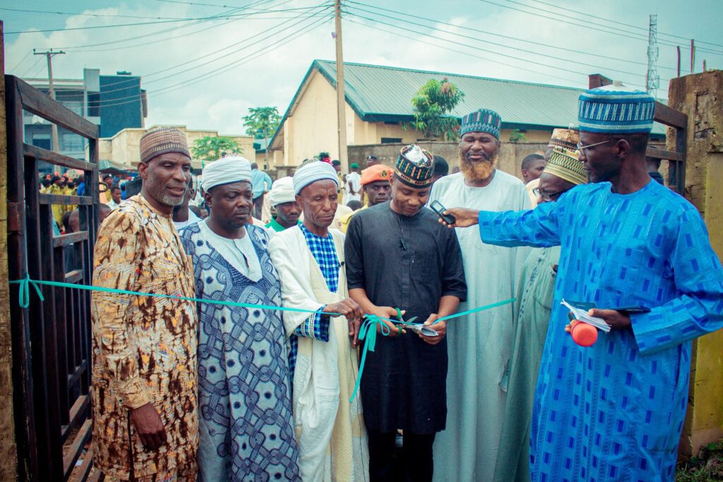 A group of men in traditional attire stand together as one man cuts a ceremonial ribbon at an outdoor Interfaith Service Project in Hayin Banki, Nigeria, with others observing and microphones present.