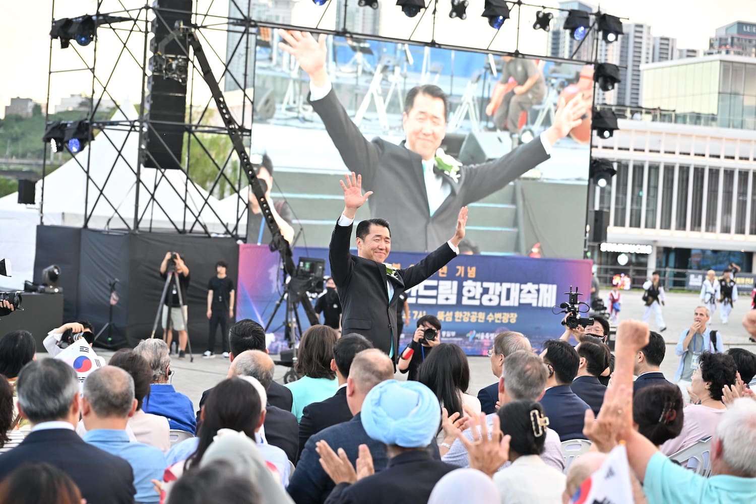A man stands and waves to an audience at Hangang Festa 2025, with a large screen behind him displaying his image as part of Korea's 80th Liberation Day celebrations.