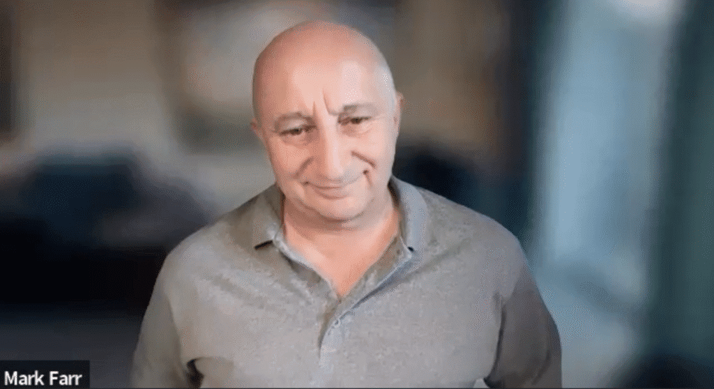 A bald man in a grey collared shirt stands indoors, smiling slightly. 
