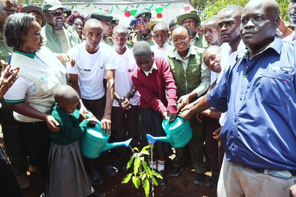 A group of students and adults water a young tree with watering cans during Global Peace Foundation Kenya’s Mazingira Day 2025, marking the national celebrations with an outdoor planting event.