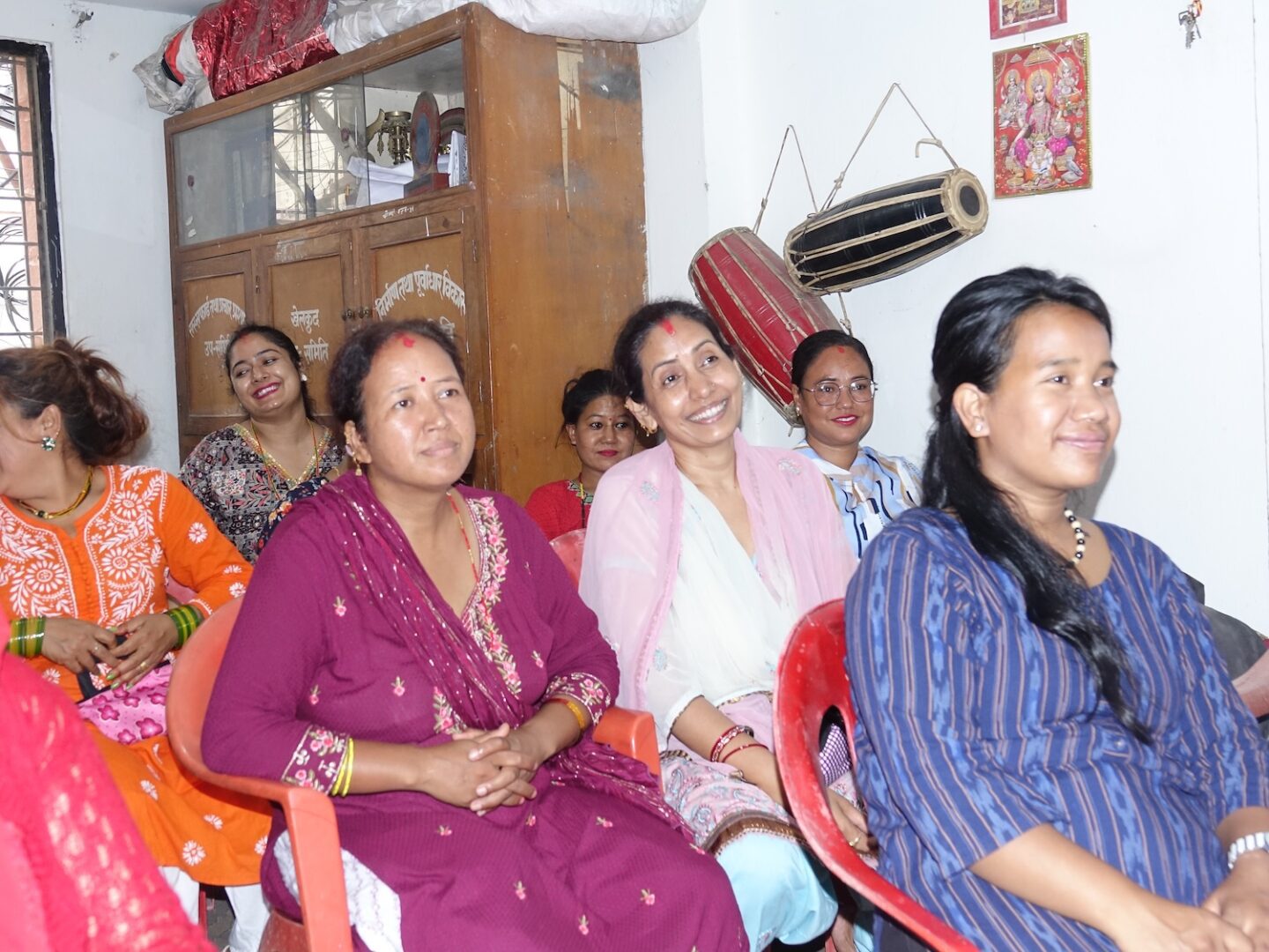 A group of women sit on red chairs indoors, smiling and wearing colorful traditional attire. Supported by GPF Nepal, they showcase economic self-reliance amid musical instruments and wall decorations in the background.