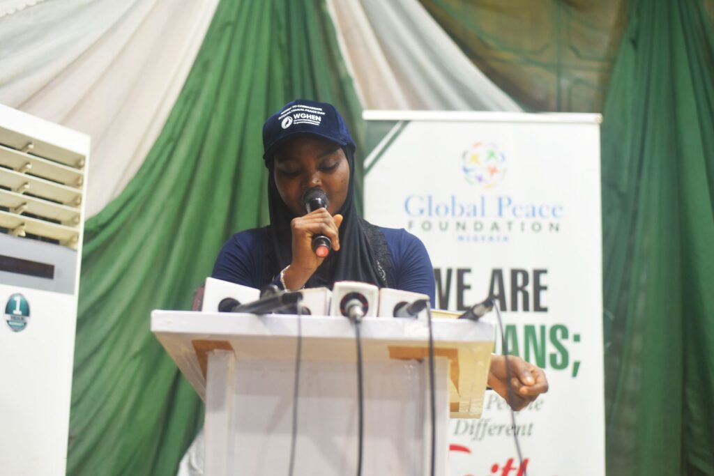 A woman speaks into a microphone at a podium during a GPF Nigeria event, highlighting community-led action. A banner and green-and-white drapery are visible in the background, marking the International Day of Peace.