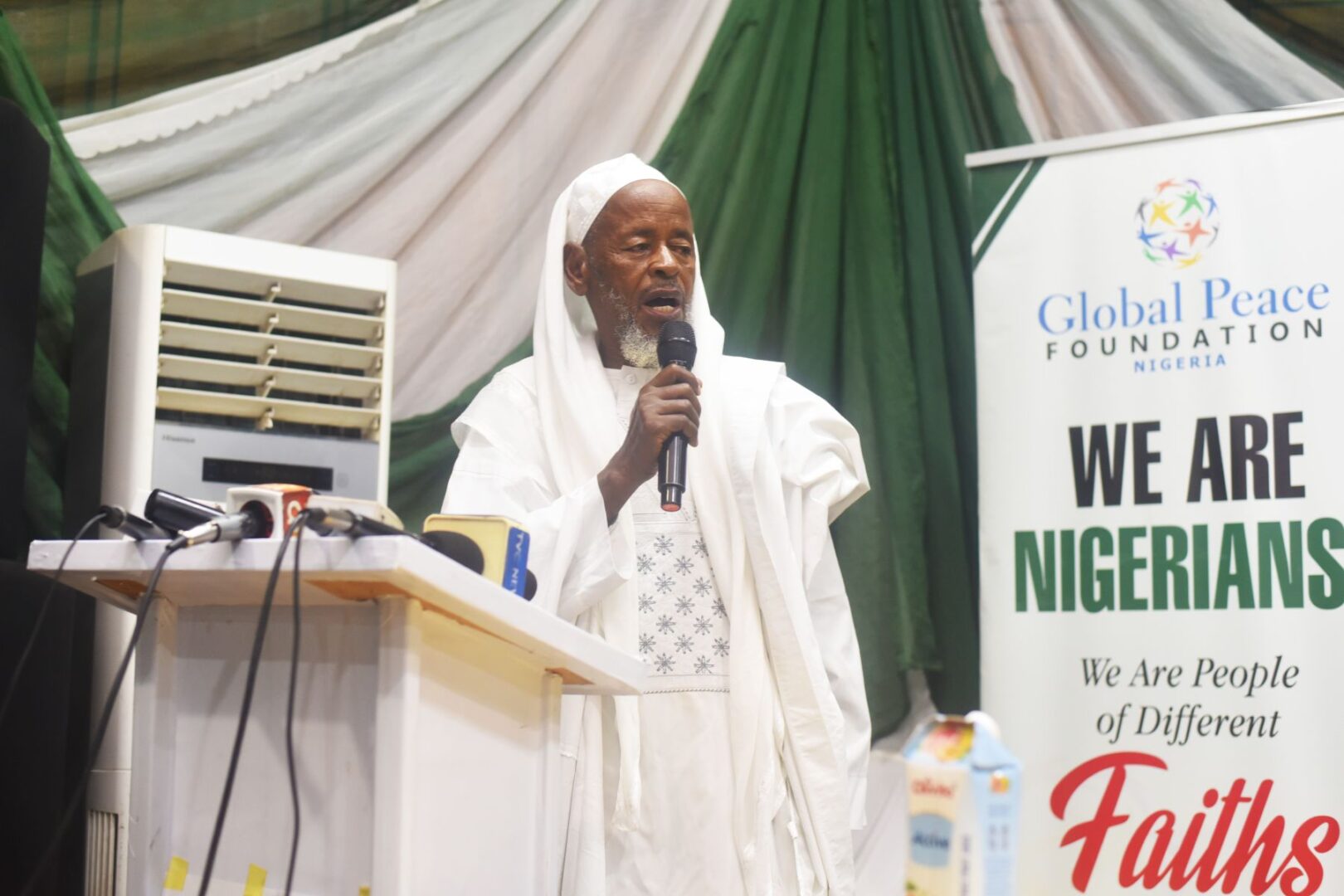 An elderly man in white traditional attire speaks into a microphone at a podium during a GPF Nigeria event, celebrating community-led action. A banner reads, "WE ARE NIGERIANS. We Are People of Different Faiths.