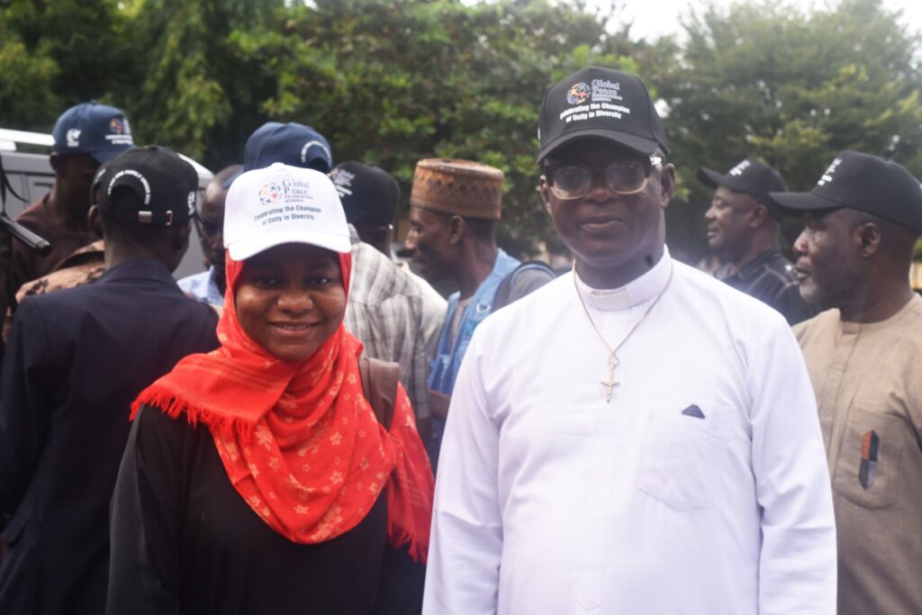 A group of people, including a woman in a red scarf and a man in white clerical clothing, stand outdoors wearing matching black and white caps during a GPF Nigeria community-led action for the International Day of Peace.