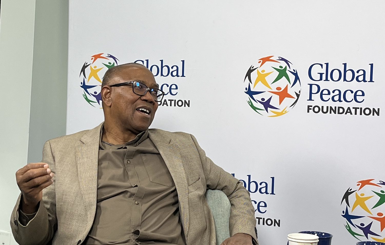 A man in a tan suit sits and gestures while speaking on ethical leadership in front of a Global Peace Foundation backdrop.