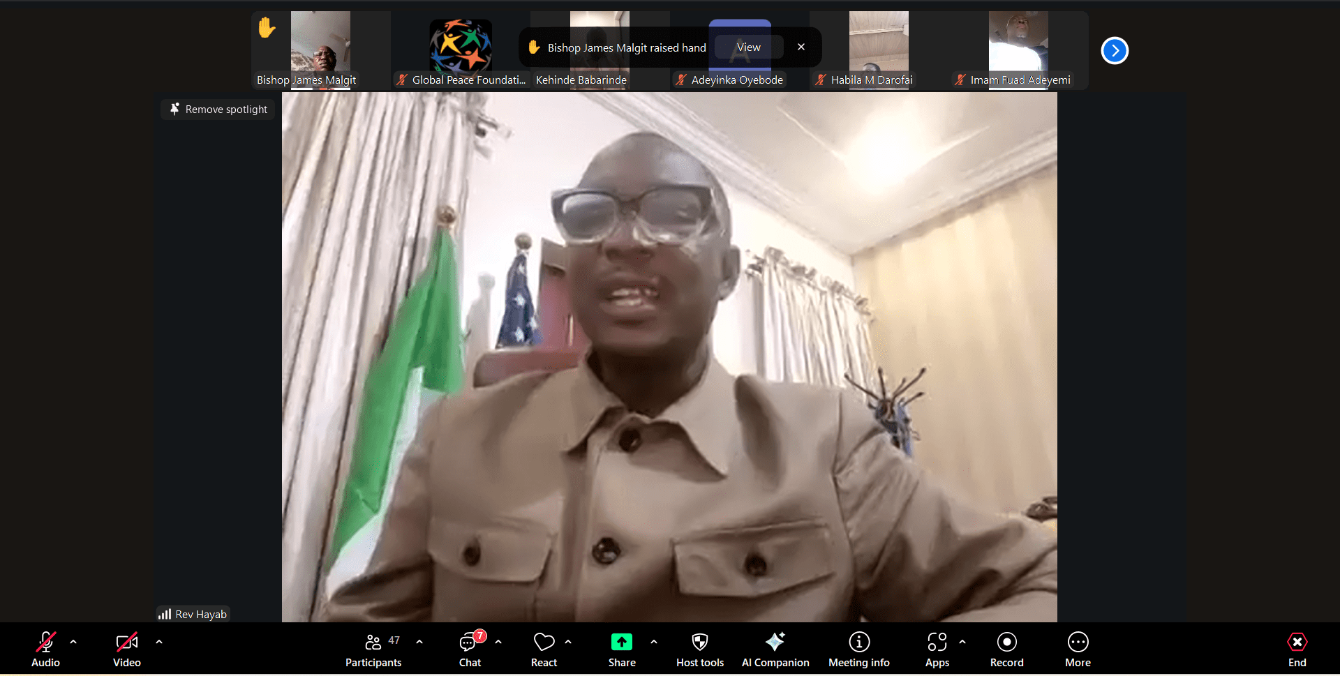 A man in glasses and a tan jacket speaks during a video conference call, discussing GPF Nigeria with community leaders. Curtains and flags are visible in the background, and Zoom interface elements appear at the top and bottom of the screen.
