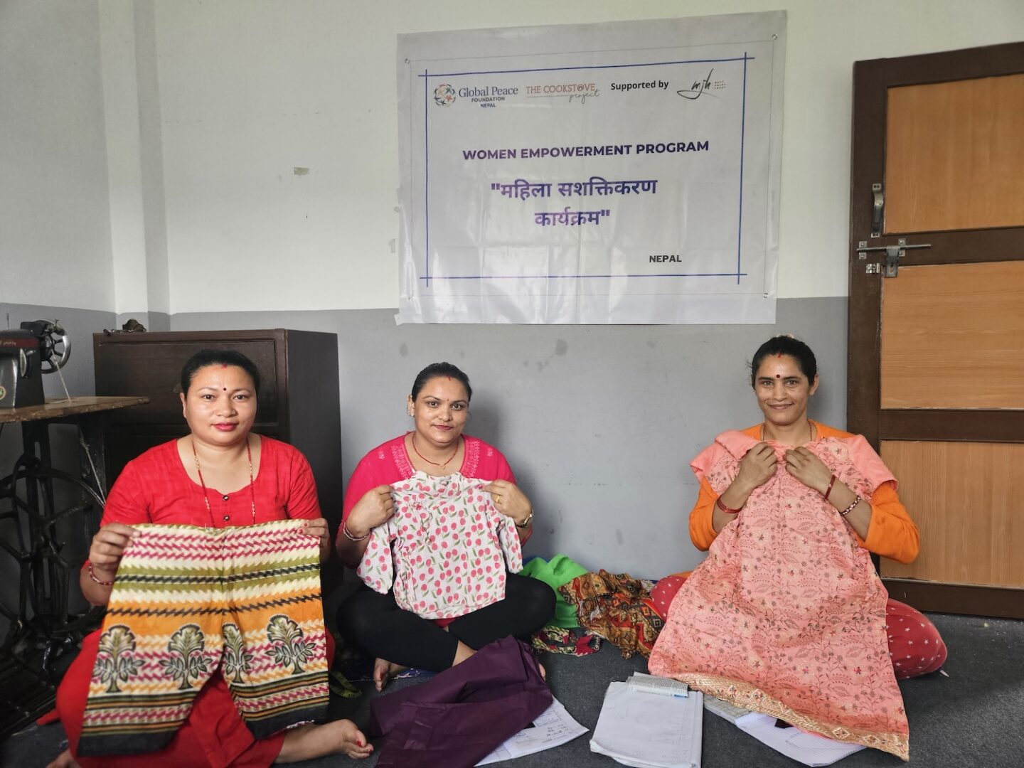 Three women sit on the floor holding handmade clothing, with documents in front of them, beneath a