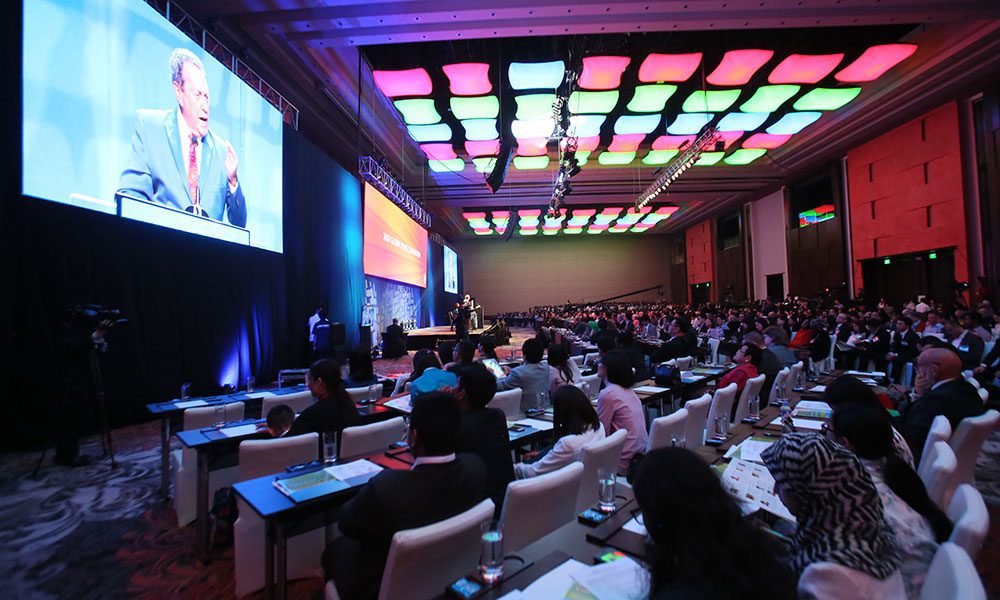 A large conference room filled with seated attendees facing a stage where a speaker presents; colorful ceiling lights illuminate the venue.