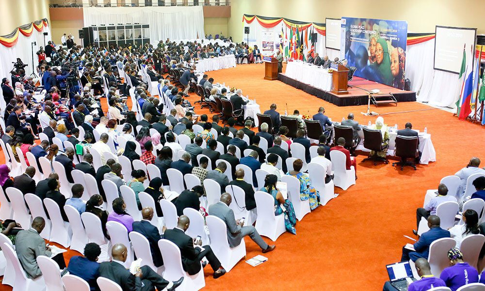 A large conference with many attendees seated in rows facing a stage with speakers; national flags are displayed along the walls.