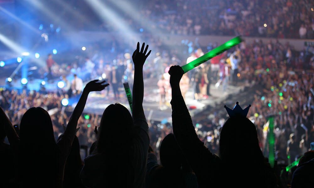 Audience members with glow sticks cheer at a crowded indoor concert, facing a brightly lit stage with performers in the background.