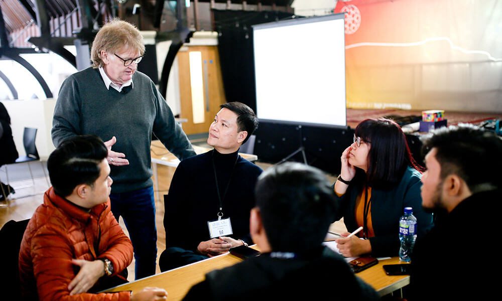 A group of people sit around a table listening to a standing man speak during a meeting or workshop in a brightly lit room.