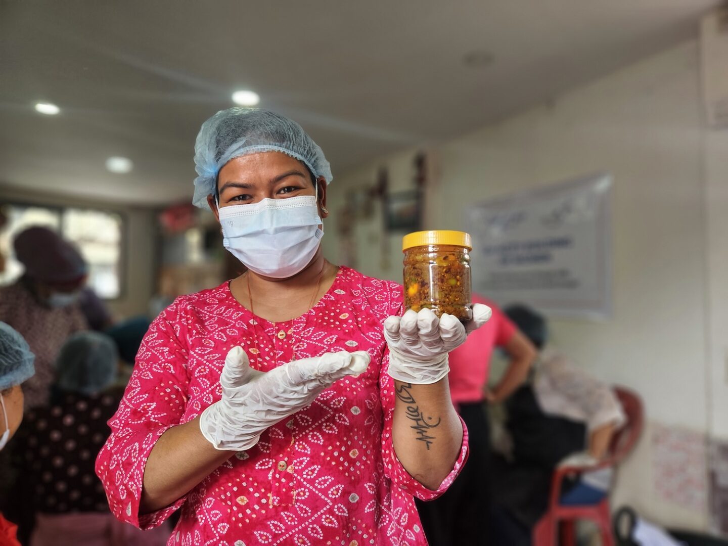 A woman wearing gloves, a hair net, and a face mask holds up a jar of preserved food, smiling inside a room with other people in the background—showcasing GPF Nepal’s efforts to empower families and promote economic self-reliance.