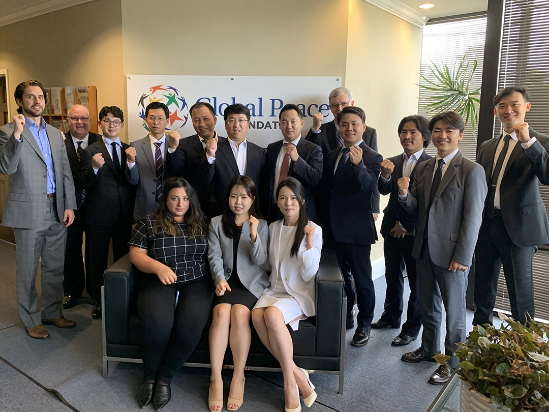 A group of people in business attire pose together in an office setting, some making a fist gesture, in front of a sign that reads “Global Peace Foundation.”.