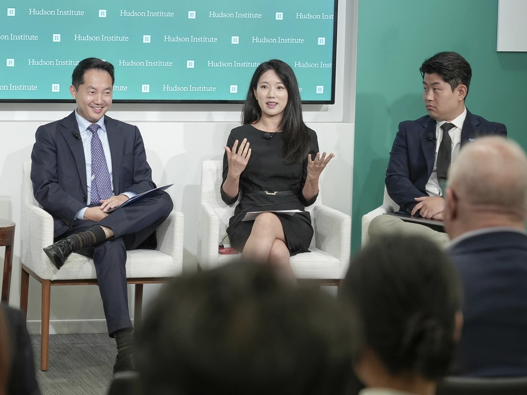 Three people in business attire sit on a panel discussion at the Hudson Institute, with two men listening as a woman speaks, while audience members watch.