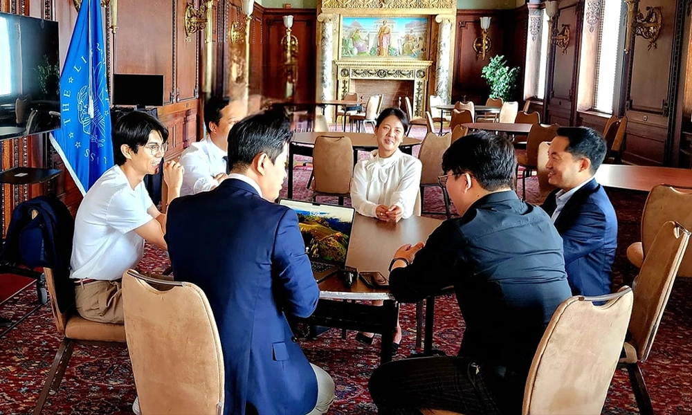 Six people sit around a table in a formal, ornately decorated room, engaged in conversation. There are flags, paintings, and wooden furniture visible in the background.