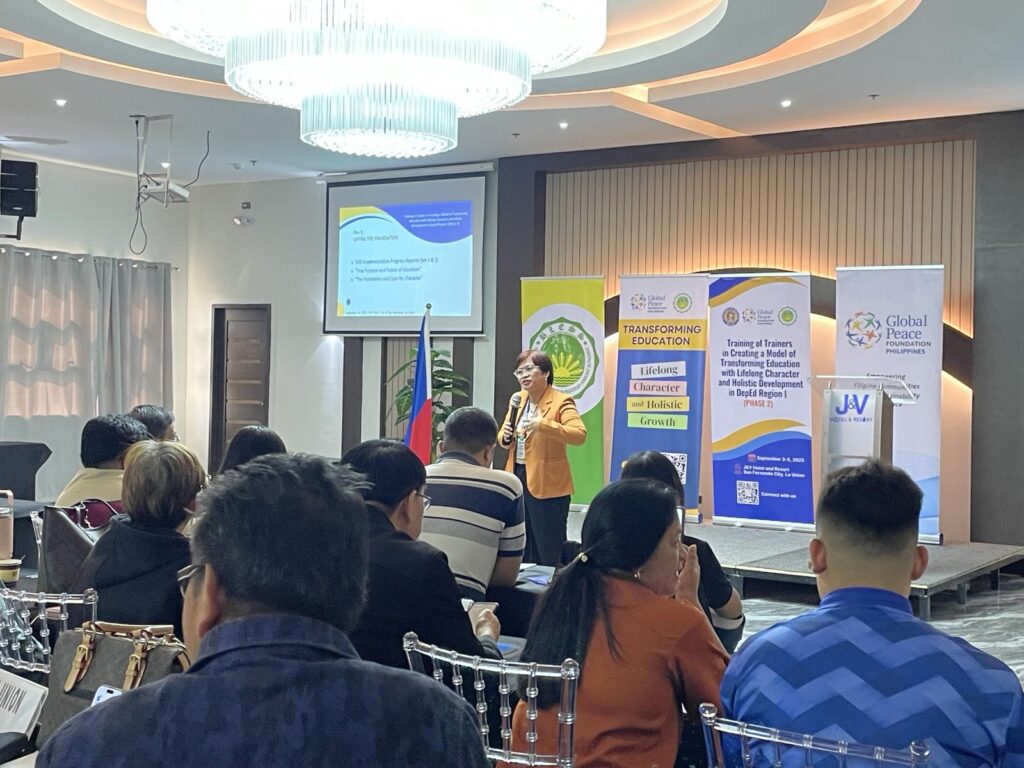 A woman speaks at a podium in front of an audience during a seminar on Transforming Education, with presentation slides and banners from the Global Peace Foundation displayed behind her.