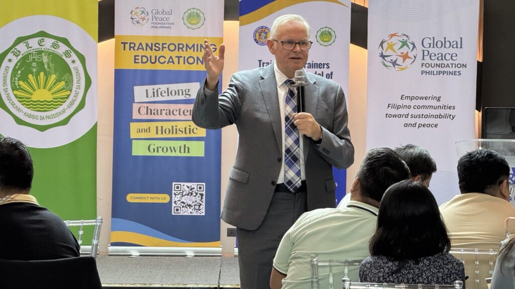 A man in a suit speaks into a microphone before seated guests, with Global Peace Foundation and Department of Education Region 1 banners about transforming education and peace displayed in the background.