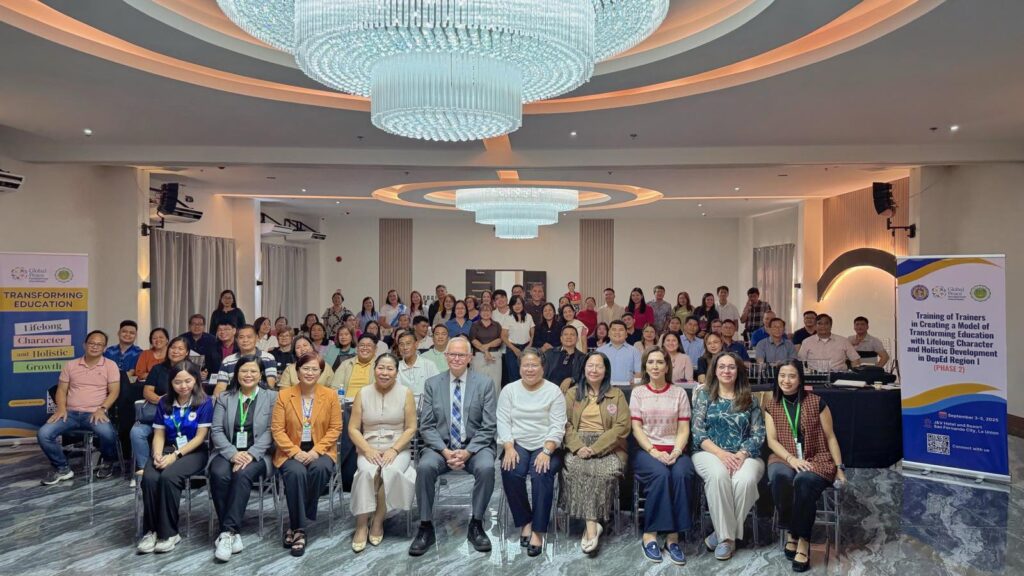 A large group poses for a photo in a conference room under chandeliers, with Global Peace Foundation and Department of Education Region 1 banners highlighting their commitment to Transforming Education, as seen on the presentation screen in the background.