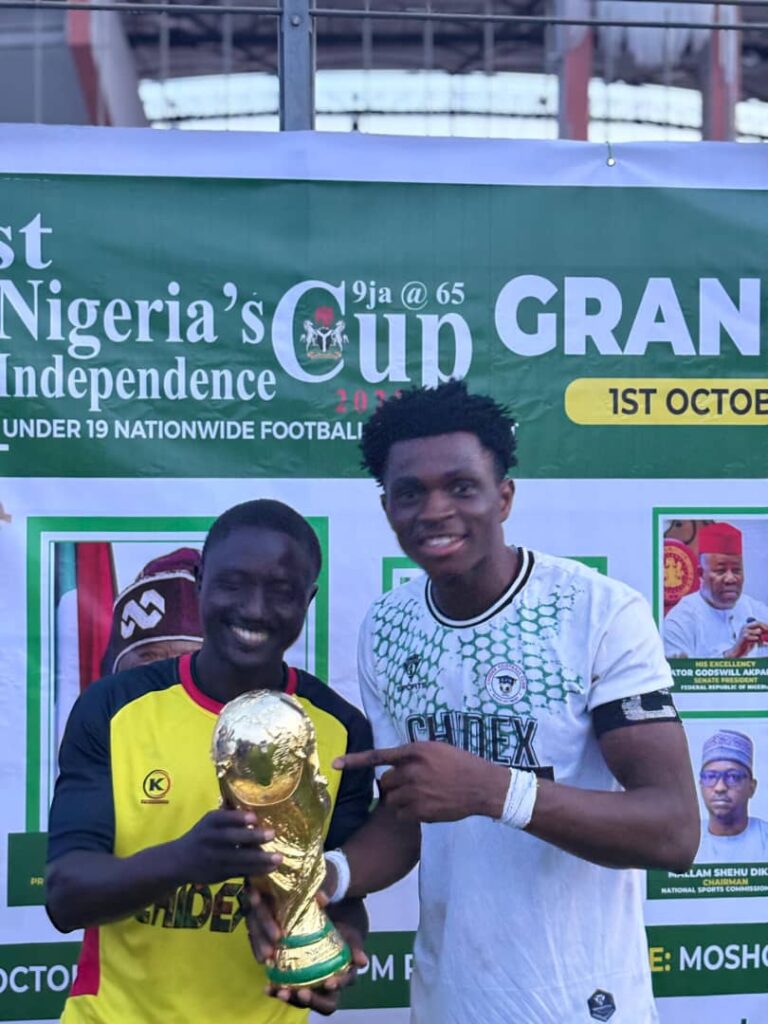 Two men standing in front of a Nigeria Independence Cup banner, smiling and holding a gold trophy together, celebrate unity and sportsmanship in Nigeria.