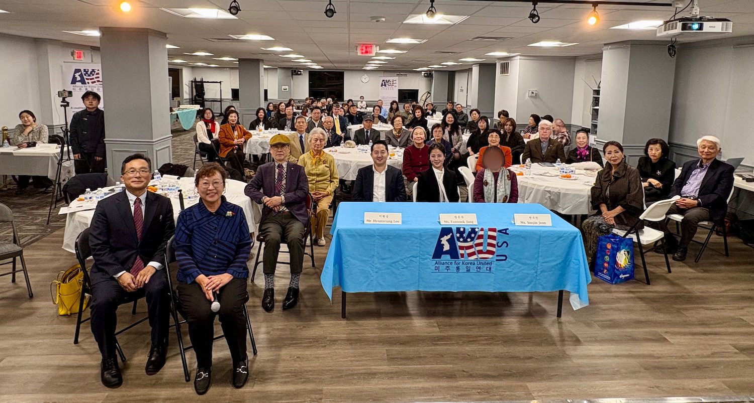 A group of people sit facing the camera in a large room with tables, attending an AKU Washington talk concert; three judges sit at a table with a blue cloth in the front center.