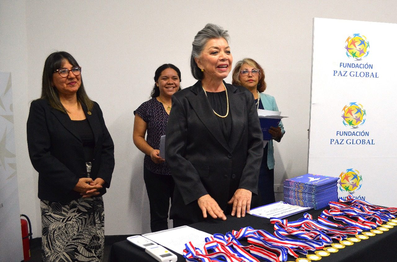 A group of women stand near a table with medals and certificates at a Fundación Paz Global event. One woman speaks, narrating peace, while others watch and smile, celebrating young voices and literary awards.