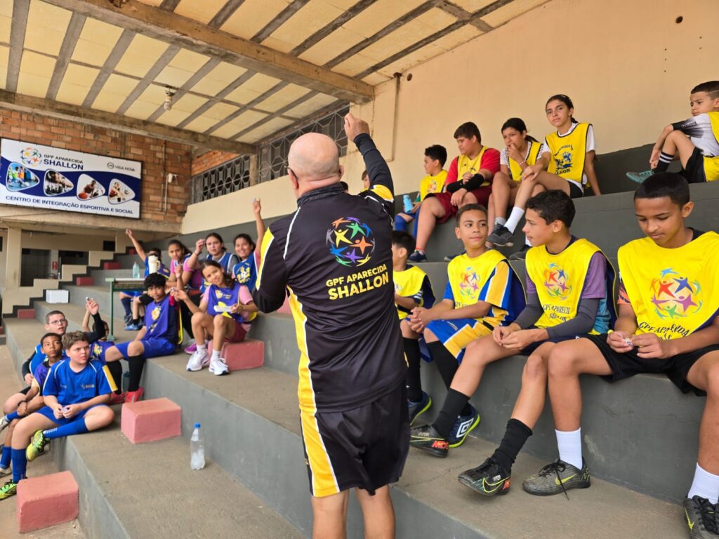 A coach in a black and yellow uniform talks to a group of children in sportswear seated on bleachers at an indoor sports facility, celebrating the spirit of sport and togetherness on Children’s Day.