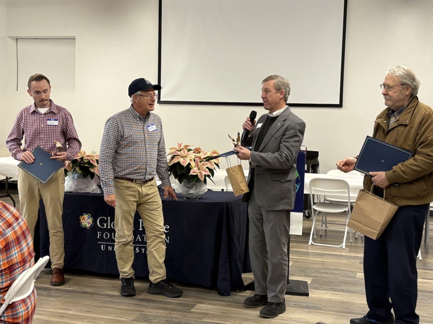 Four men stand near a table with poinsettias; one man speaks into a microphone while handing a certificate and gift bag to another, celebrating achievements in the Montana grassroots project and fostering cross-cultural dialogue.