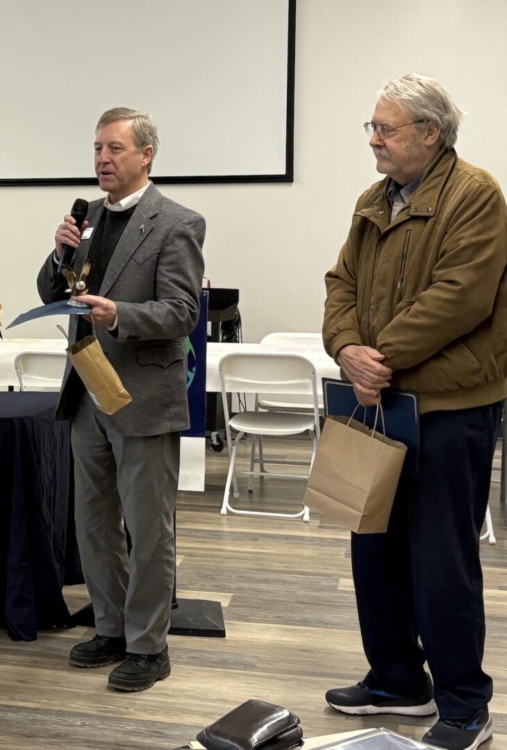 Two men stand indoors at the Global Peace Forum in Montana; one speaks into a microphone holding a small award and gift bag, while the other holds a certificate and gift bag, both facing an audience.