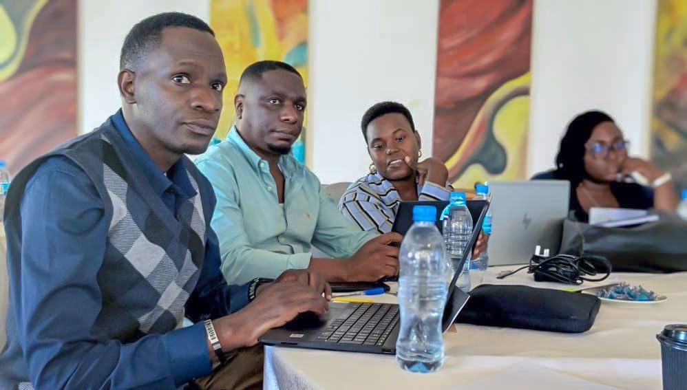 Four people sit at a table with laptops and bottled water, appearing focused and engaged in a shared learning session on local peace structures, possibly organized by GPF Tanzania.